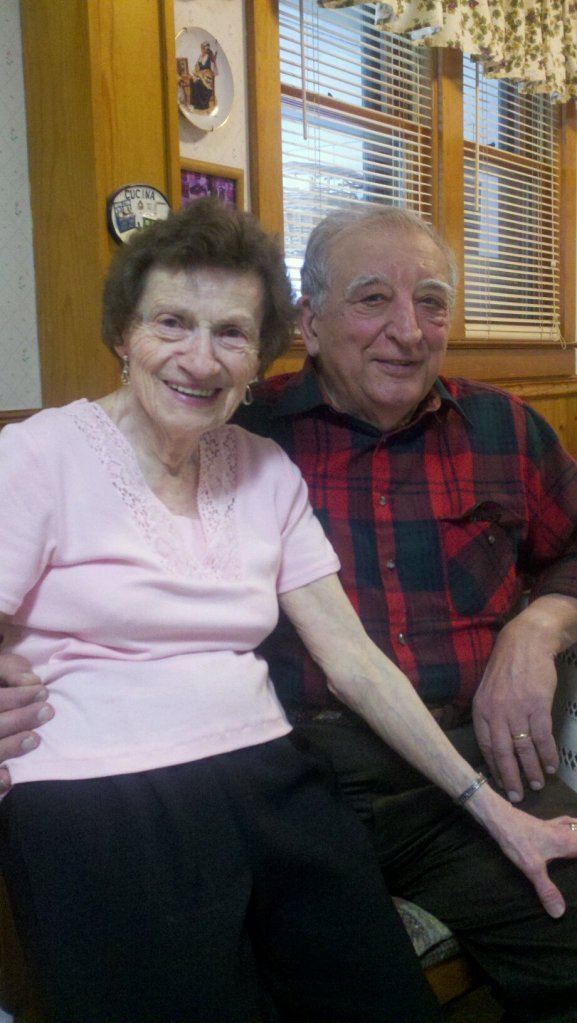 An older woman is sitting on the knee of an older gentleman. Both are smiling. She has brown hair and is wearing a pink shirt. He is balding, with white hair, and is wearing a red and green plaid shirt.