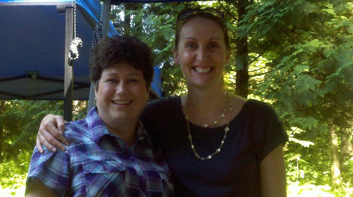 Photo of two women standing with arms around each other. The woman on the left has short dark hair and is wearing a plaid shirt. The woman on the right has blond hair pulled back in a ponytail and is wearing a blue tee-shirt and a necklace.