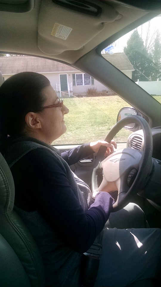 Photo of the profile of a woman seated behind the steering wheel of a car. She has brown hair pulled back in a pony tail and is wearing glasses.