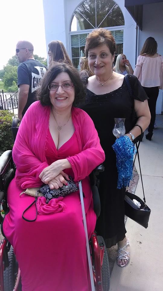 A woman seated in a wheelchair wearing a long pink dress and a pink shawl sits next to a woman wearing a black dress. The woman in the wheelchair has brown hair and is wearing glasses. She has a camera on her lap. The woman standing next to her is holding a glass and a blue shawl.